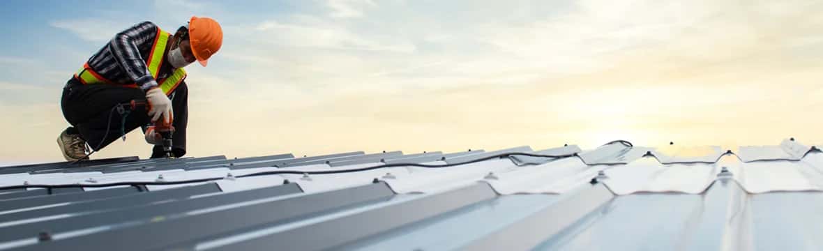 Roofer installing metal roof panels with power drill at sunset