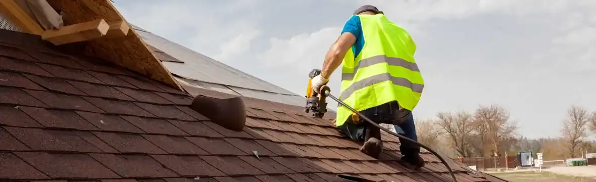 Roofer installing brown asphalt shingles with nail gun on home